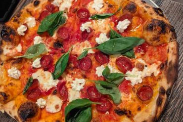 Pizza topped with fresh basil, cherry tomatoes, mozzarella cheese, and pepperoni slices on a wooden table centered in the image.