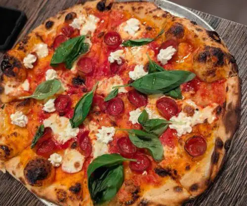 Pizza topped with fresh basil, cherry tomatoes, mozzarella cheese, and pepperoni slices on a wooden table centered in the image.
