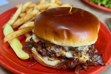 Slow cooker BBQ pulled pork sandwich with melted cheese on a toasted bun, served with crispy fries and pickles on a red plate at a casual American restaurant.