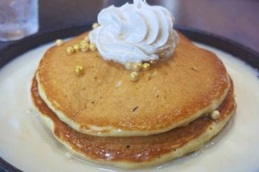 Fluffy stack of pancakes topped with whipped cream and gold sugar pearls, served with vanilla syrup, ideal for breakfast or brunch in a cozy cafe setting.