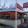 Vintage burger stand with red and silver exterior at dusk, featuring "We are OPEN" sign, neon lights, and signage advertising classic American meals like hamburgers and French fries.