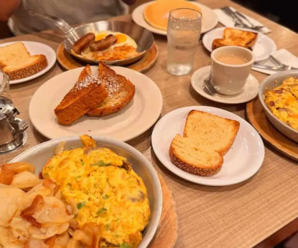 Crispy French toast with powdered sugar and breakfast sides, including scrambled eggs, sausage, toast, and coffee, served on a rustic wooden table in a cozy café setting.