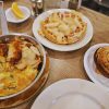 Warm breakfast with scrambled eggs, a fruit waffle topped with pear slices, and a cinnamon roll on white plates at a cozy cafe. Lemon wedges and coffee add to the breakfast spread.