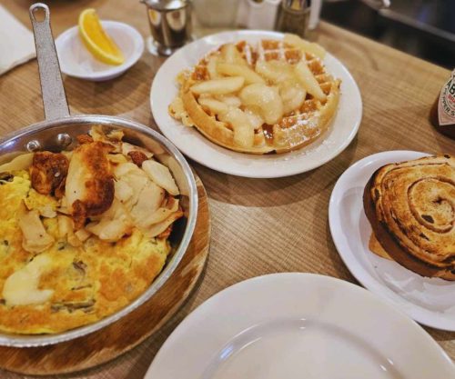 Warm breakfast with scrambled eggs, a fruit waffle topped with pear slices, and a cinnamon roll on white plates at a cozy cafe. Lemon wedges and coffee add to the breakfast spread.