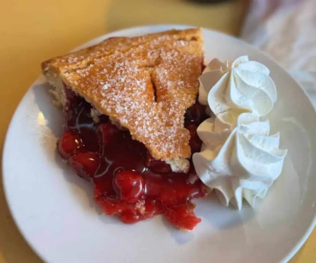 Delicious cherry pie with a golden crust, topped with powdered sugar, served with whipped cream on a white plate. Perfect for dessert or a sweet treat at any destination.
