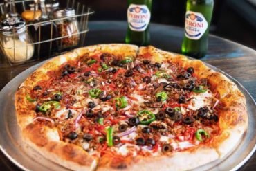 Freshly baked pepperoni pizza topped with black olives, green peppers, and onions, served on a metal tray with cold Peroni beers in the background; perfect for a casual dining experience.