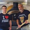 Authentic pizza oven with two smiling men holding pizza tools in a cozy pizzeria kitchen, promoting delicious Italian pizza and culinary travel experiences.