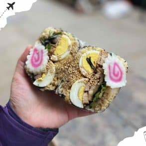 Sushi roll with boiled eggs, green herbs, and sesame seeds held in hand, representing Japanese cuisine and travel food experiences.